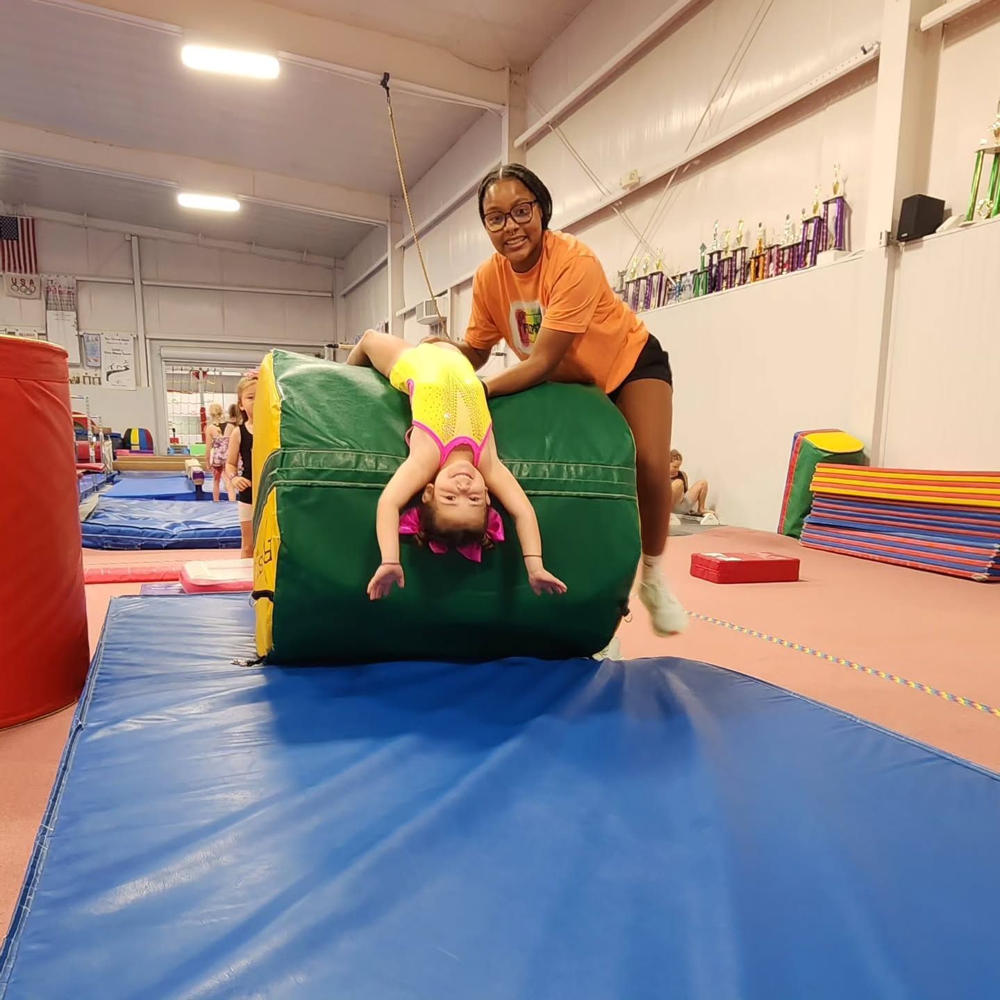 Child tumbling on a green foam block with a coach in an indoor gym, on blue mats.
