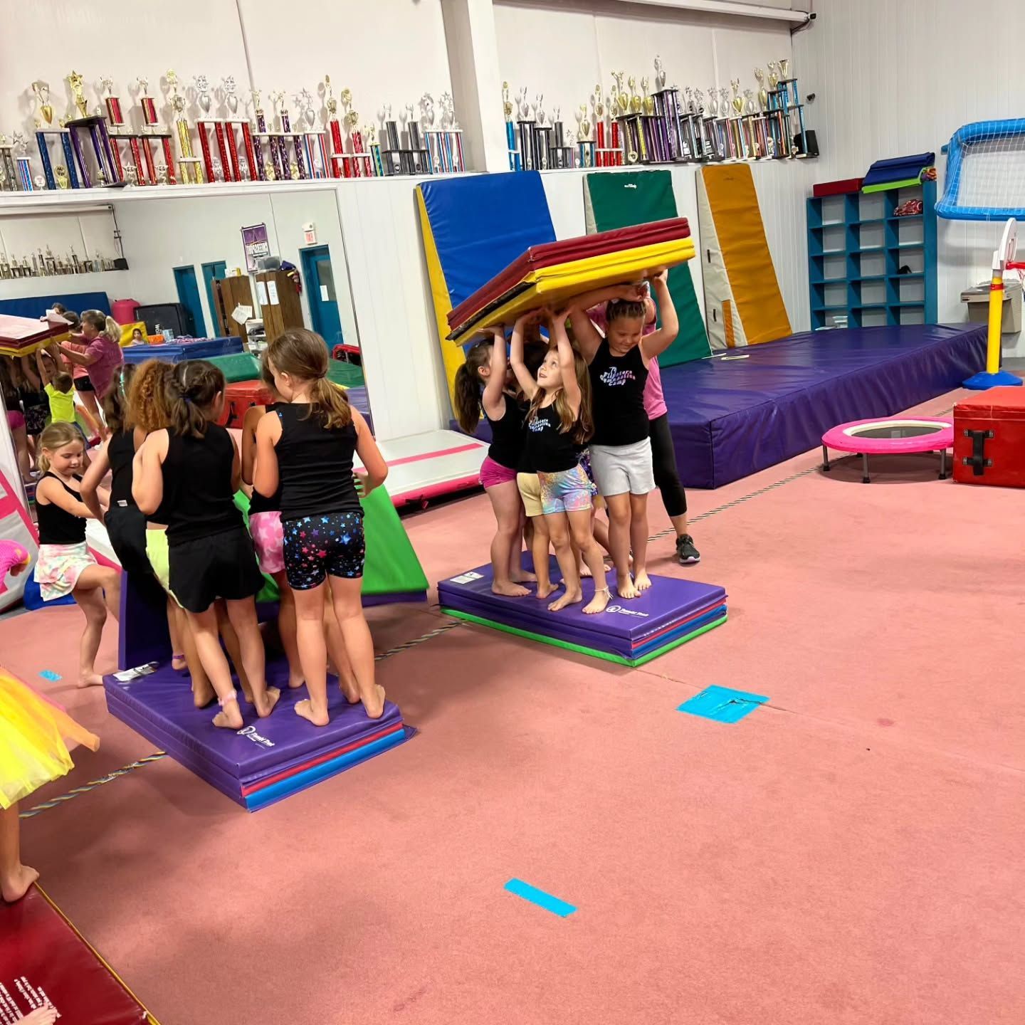Children practicing gymnastics on balance mats in a colorful gym, holding a foam mat together overhead.