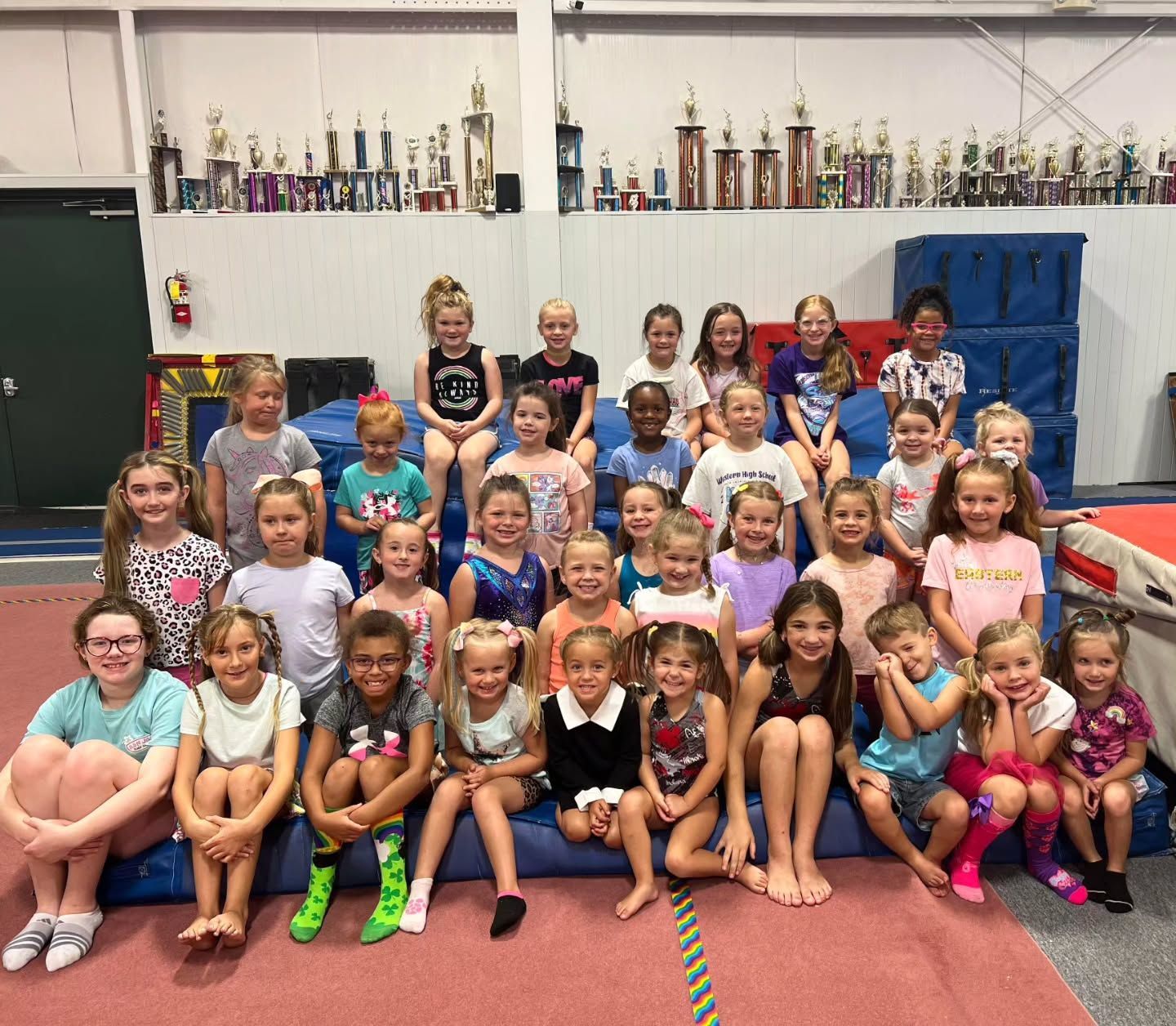 Group of children and adults posing in a gymnasium with trophies displayed on shelves behind them.