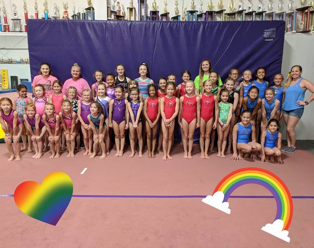 Large group of gymnasts in colorful leotards pose indoors with rainbow heart and rainbow graphics.