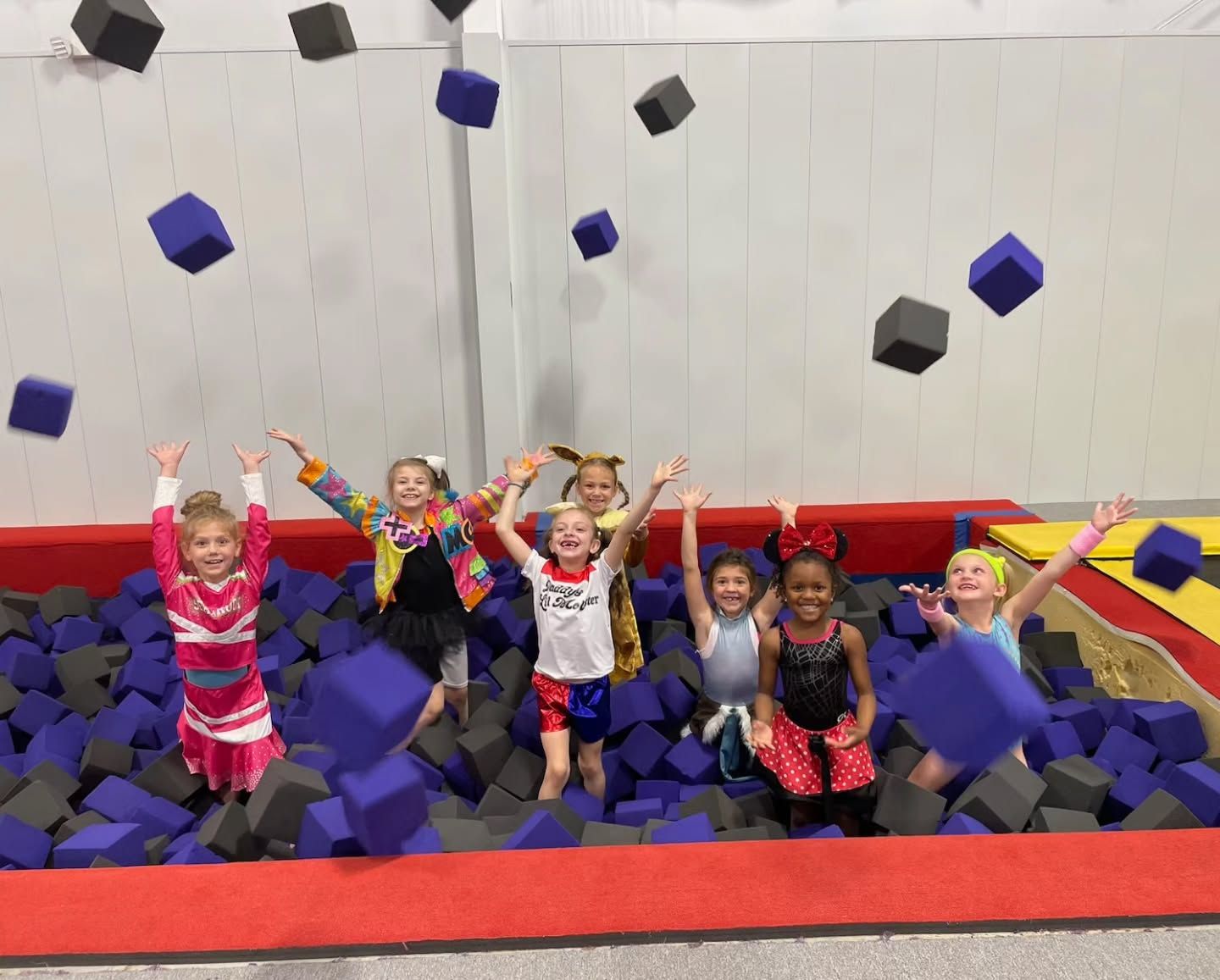 Children jumping in a foam pit, with blue and black cubes flying around a trampoline gym.