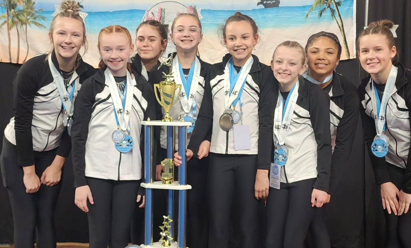Seven smiling athletes in medals stand behind a trophy with a beach mural backdrop.