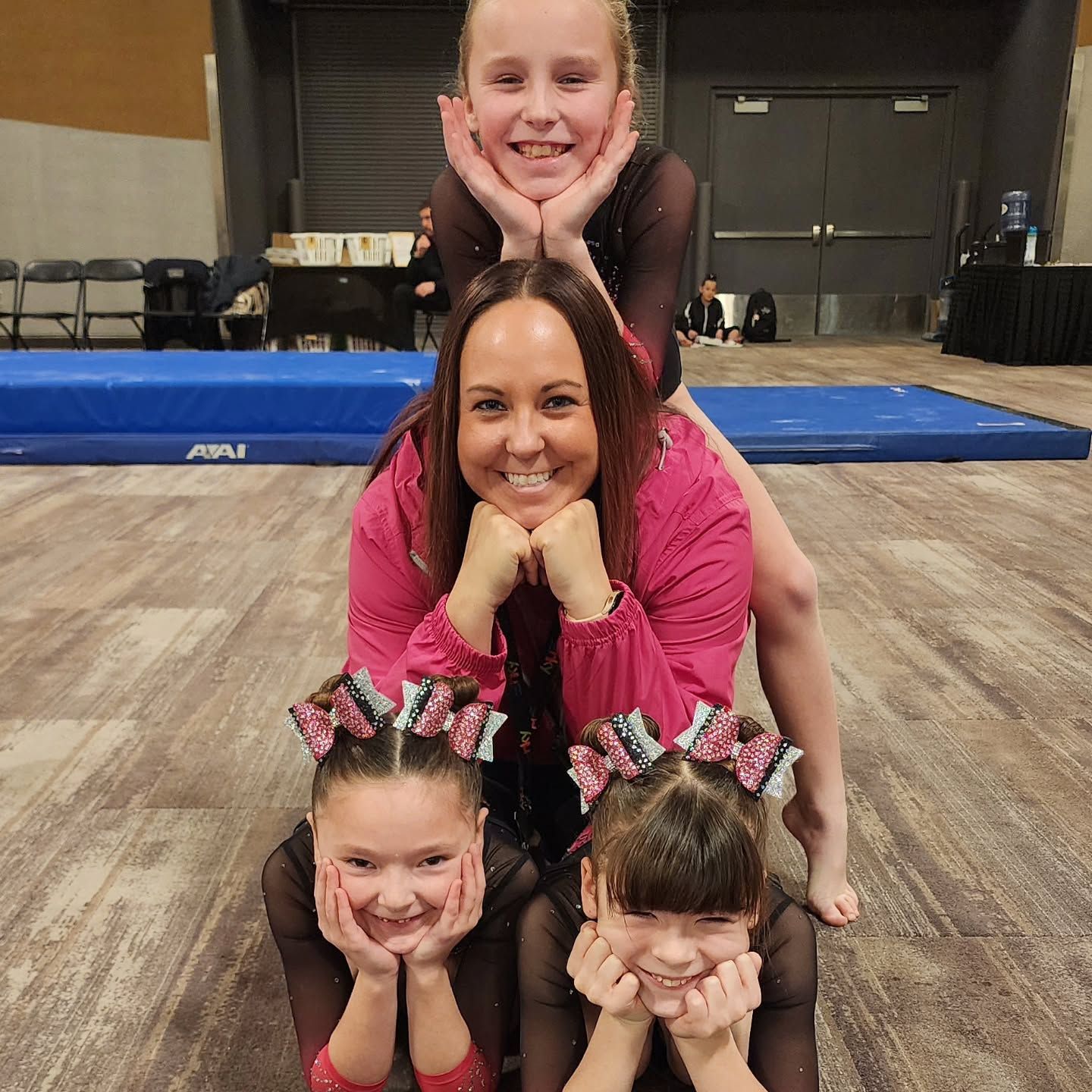 Four people stacked in a playful pose on a gym floor, smiling with hands on their chins.