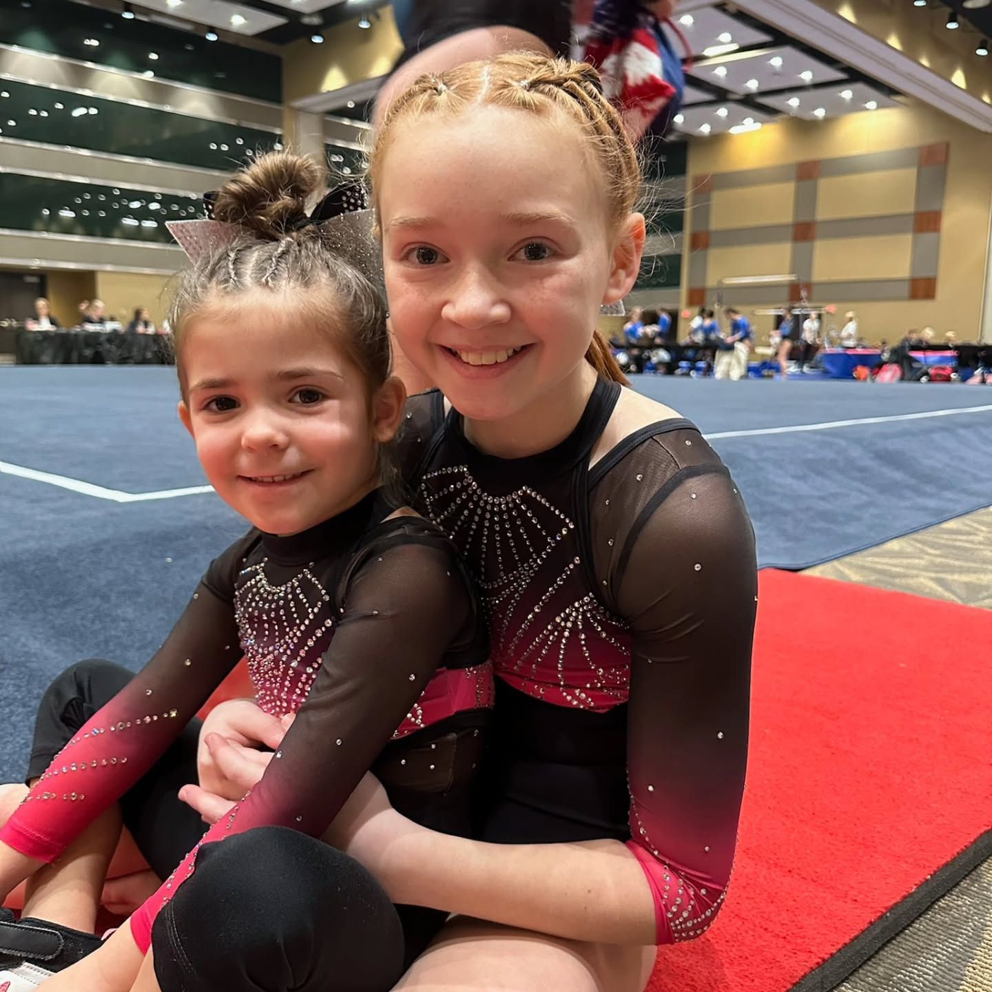 Two gymnasts in black and pink leotards sit smiling on a red mat in an indoor arena.