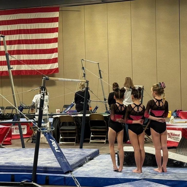 Gymnastics team watching a routine on uneven bars in a competition venue with an American flag backdrop