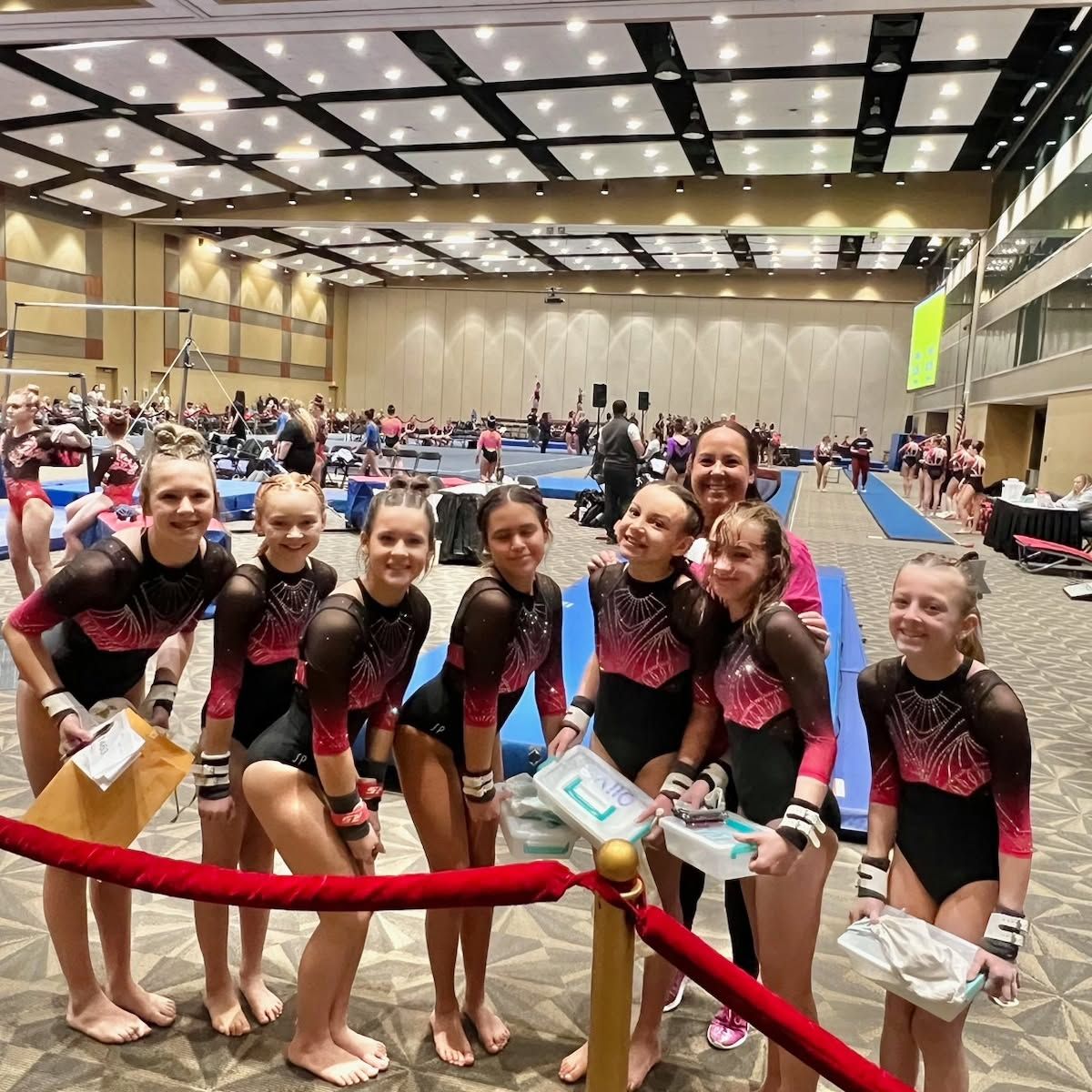 Gymnasts in matching black and pink leotards pose behind a red rope at a competition venue.