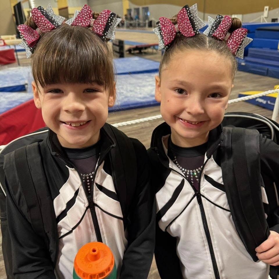 Two young gymnasts smiling in matching black and white uniforms with pink bows, standing in a gym.