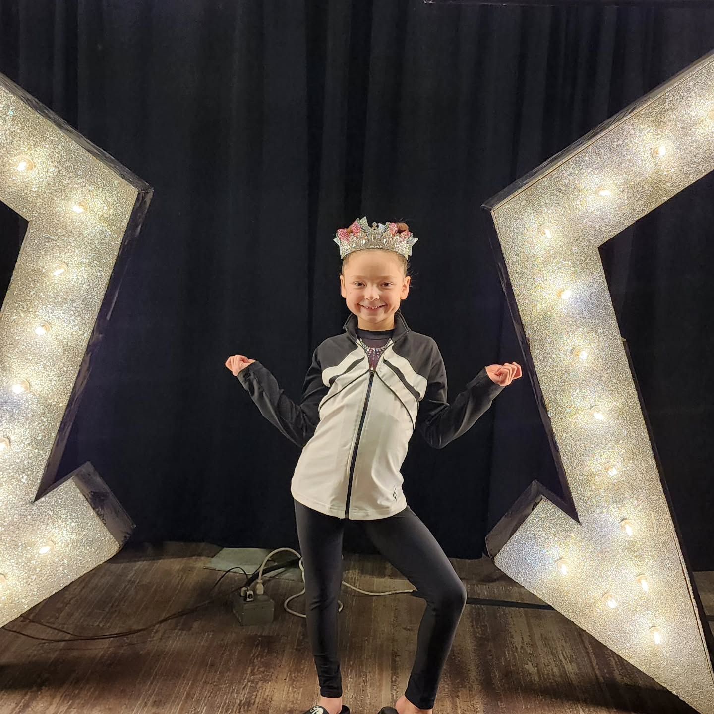 Smiling child posing on a stage between two large illuminated star props, wearing a black-and-white outfit.