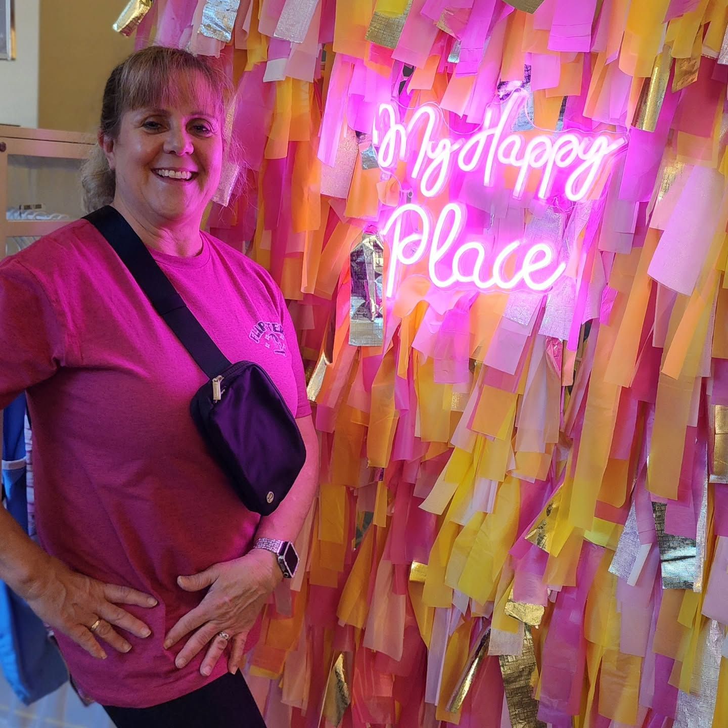 Woman in a pink shirt standing beside a neon “My Happy Place” sign on a pink and yellow paper backdrop