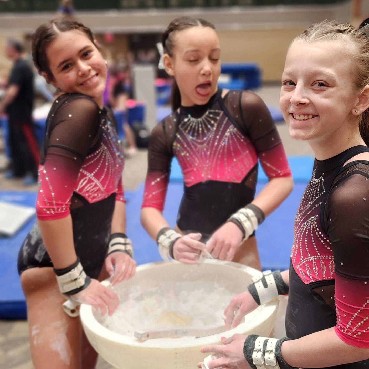 Three gymnasts in pink and black leotards smiling around a chalk bowl in a gym