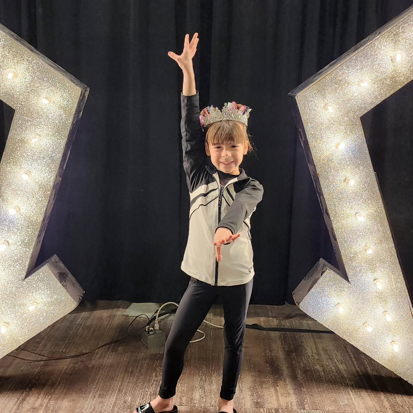Child posing on a stage with one arm raised, wearing a crown, between two lit decorative panels.
