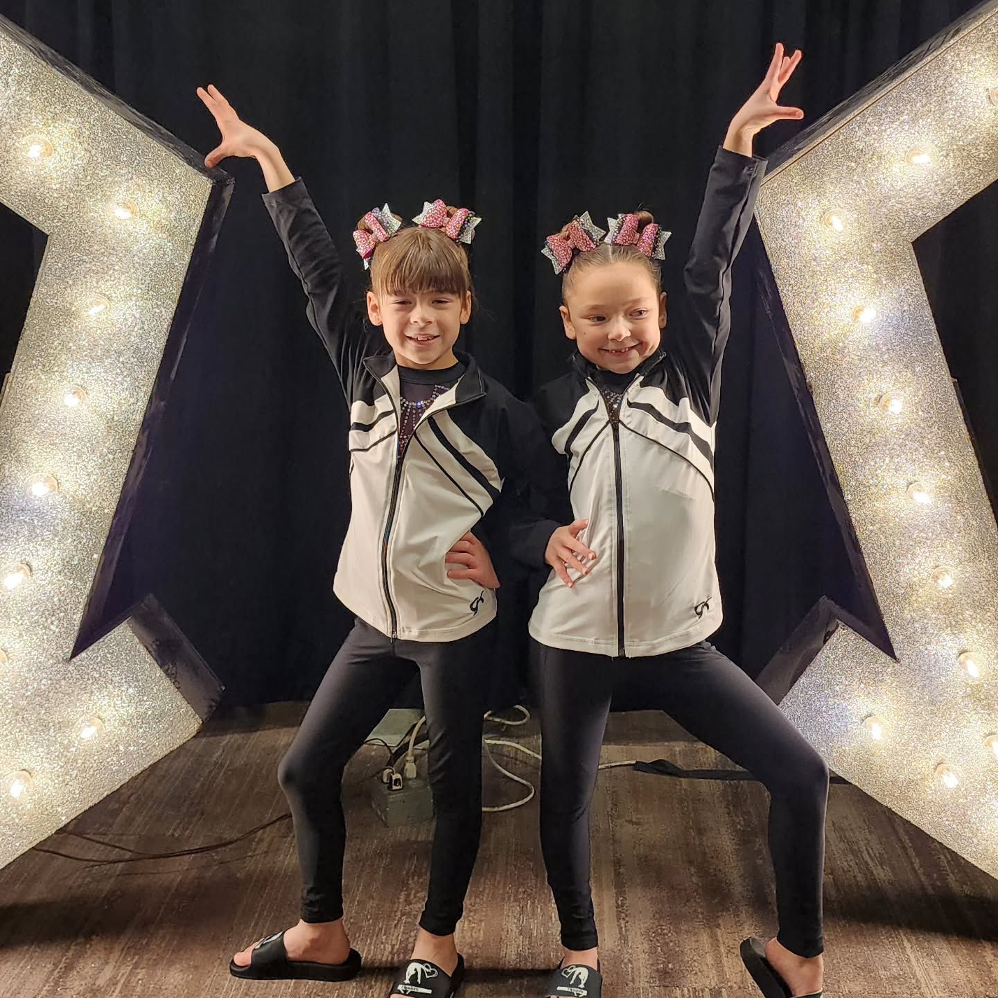 Two children posing on a lit stage in black-and-white outfits with arms raised in a playful dance pose