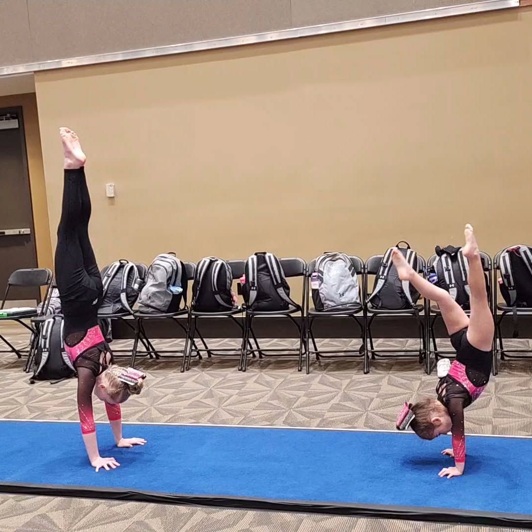 Two gymnasts in pink leotards perform handstands on a blue mat in a gym hall.