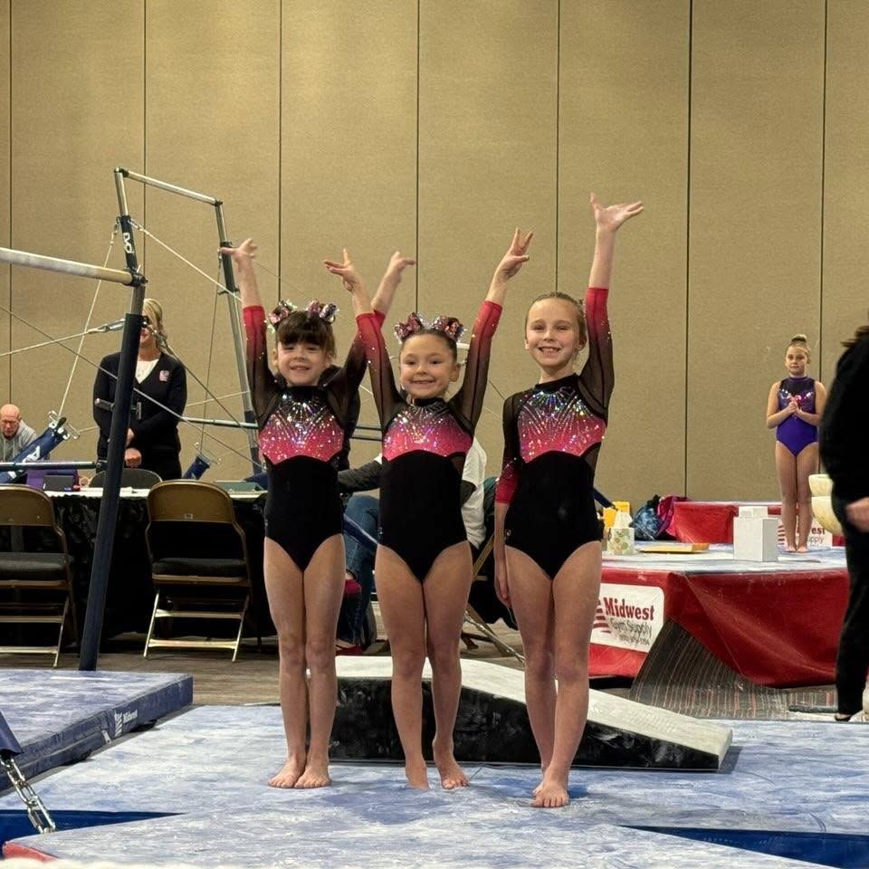 Three young gymnasts in black and pink leotards pose with raised arms on a competition mat.