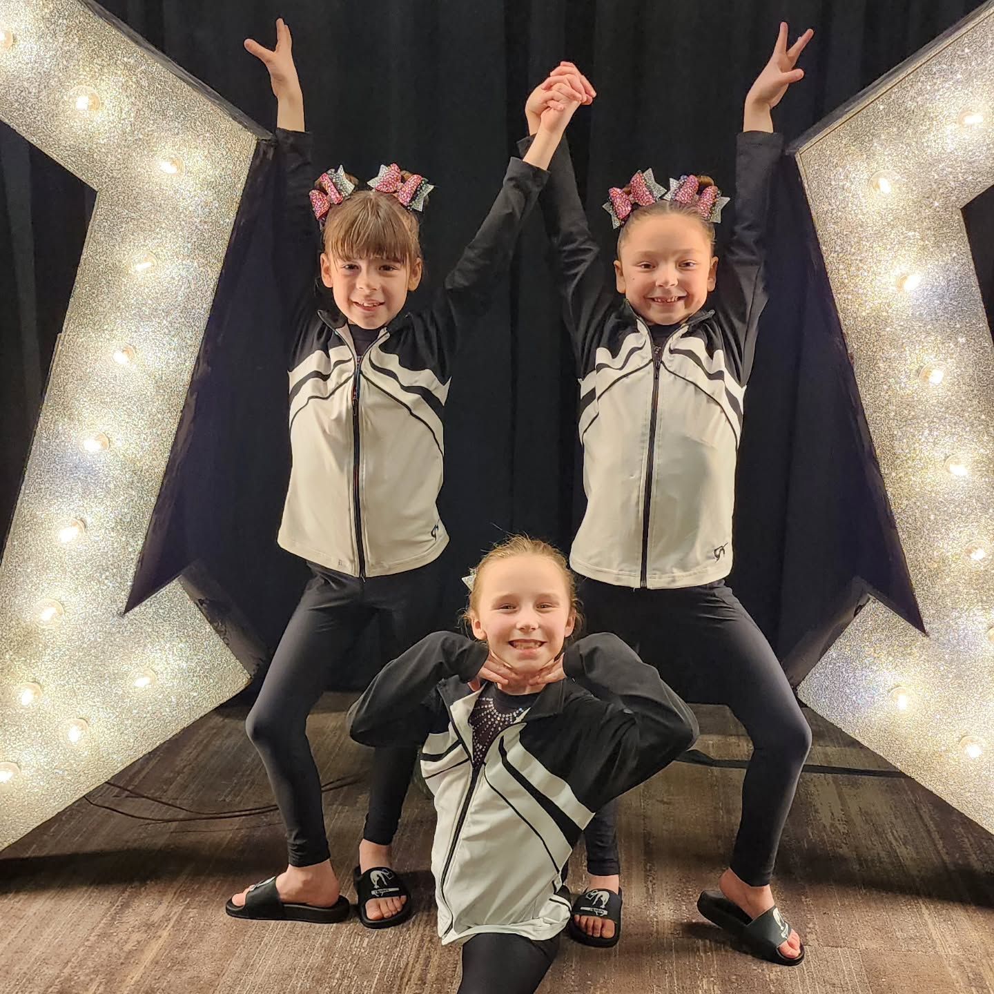 Three children in matching black-and-white outfits posing with arms raised in front of a glittering star backdrop