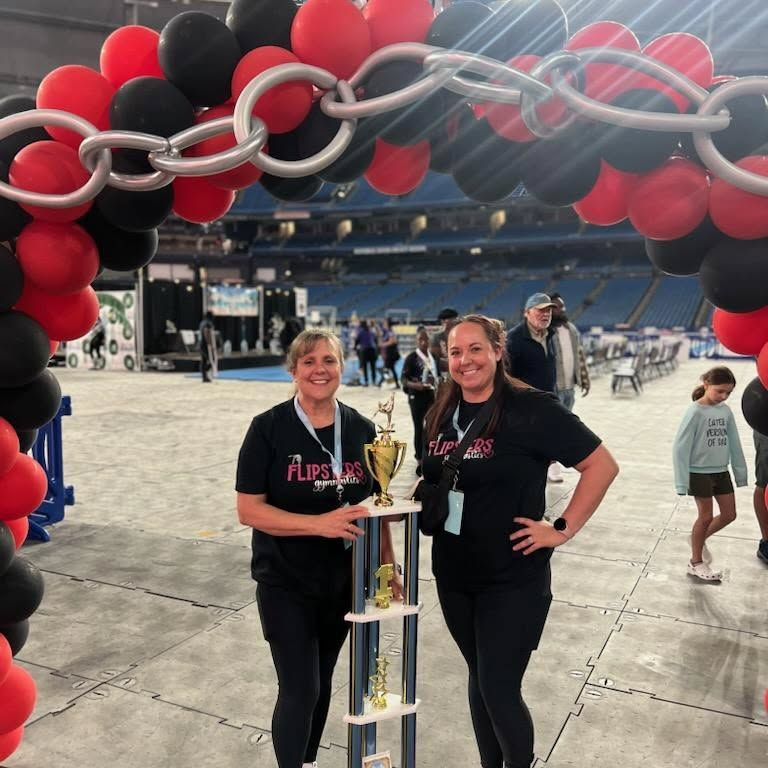 Two women posing with a trophy under a red-and-black balloon arch at a sports event.