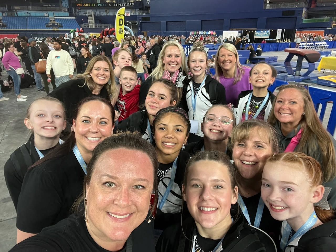 Group smiling for a selfie in a stadium, with blue seats and event tents in the background.