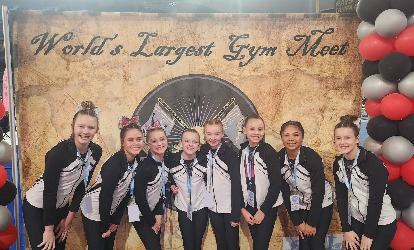 Seven gymnasts posing in front of a Worlds banner and balloon arch, wearing black-and-white uniforms
