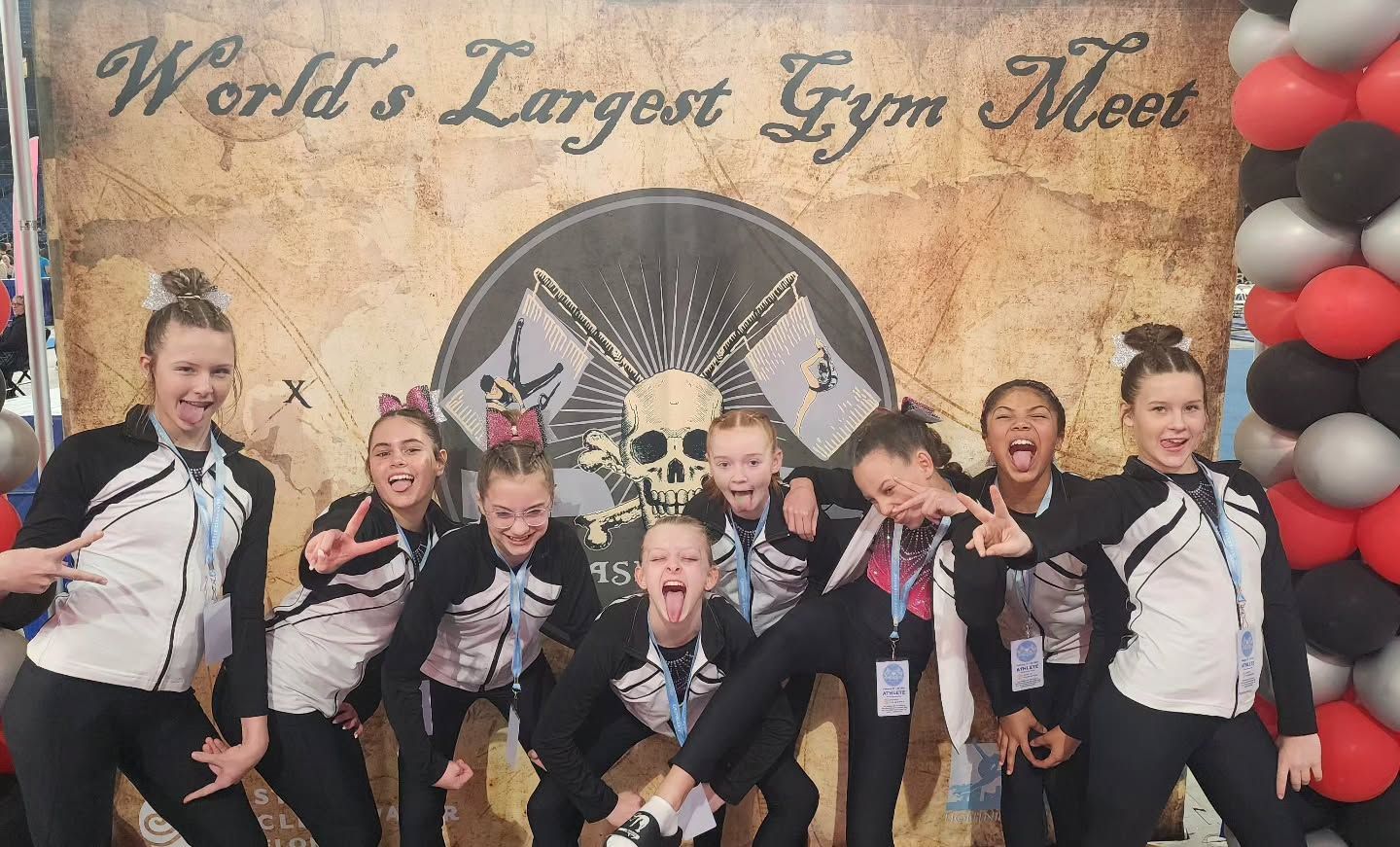 Group of gymnasts posing and cheering in front of a “World’s Largest Gym Meet” backdrop