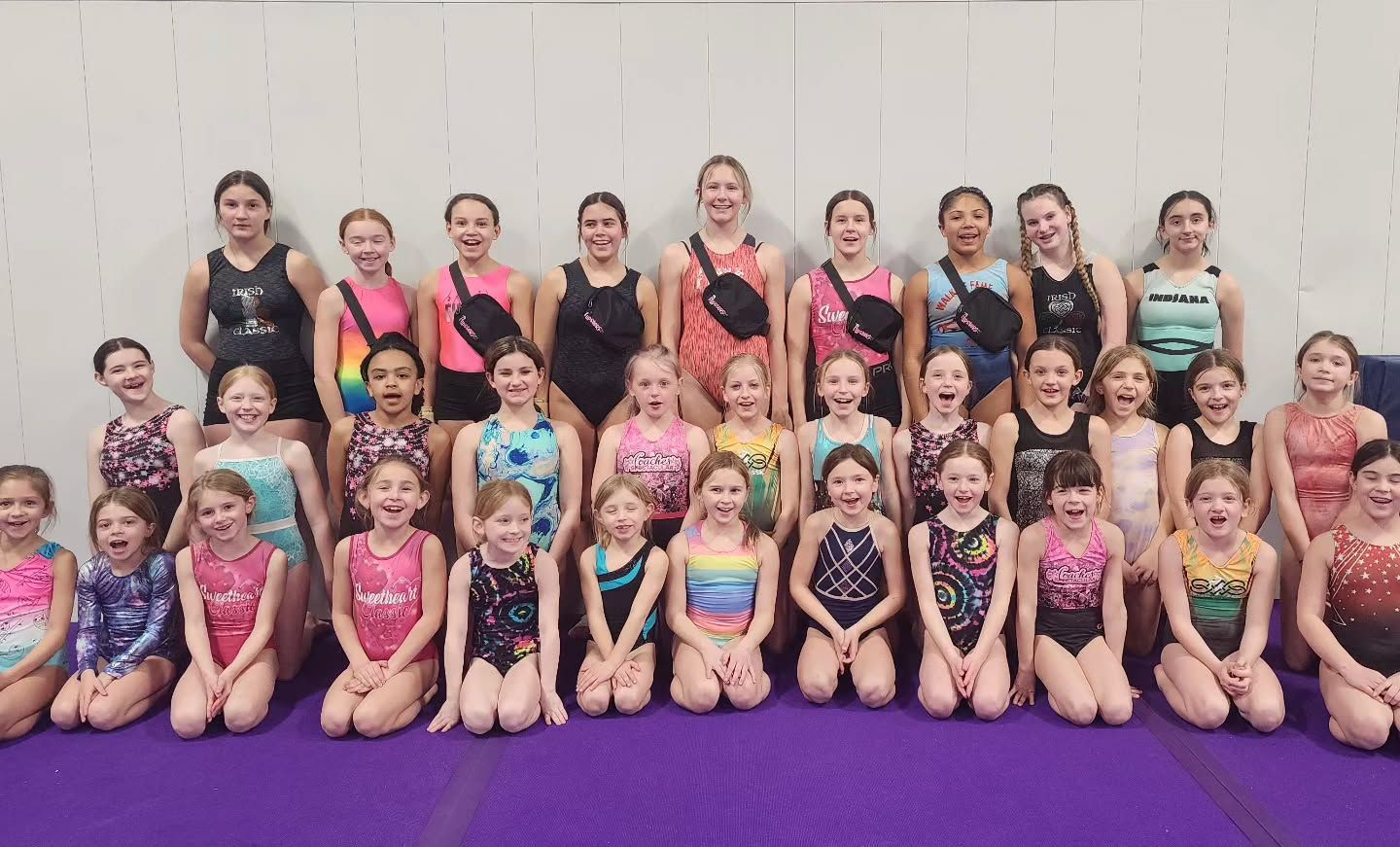 Large group of children and coaches posing in sparkly dance costumes on a purple floor against a wall
