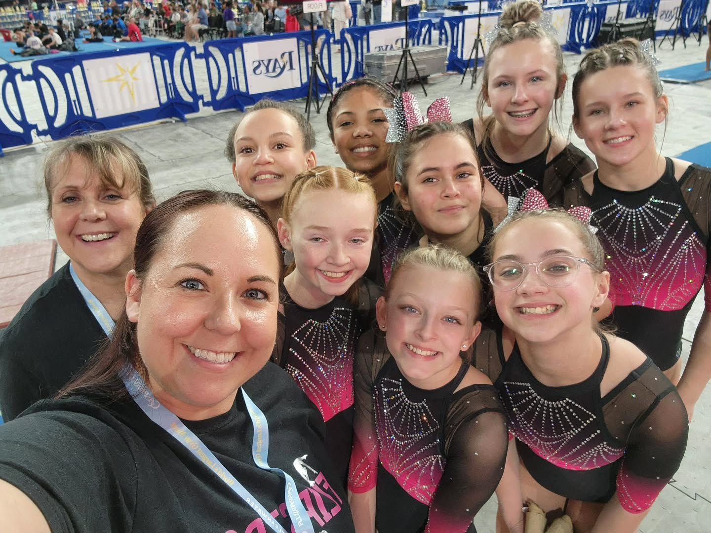 Group selfie of nine gymnasts in sparkly black and pink leotards at a gymnastics meet