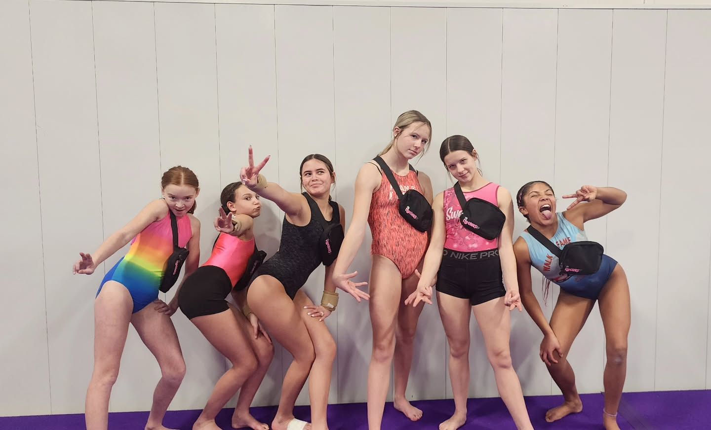 Six girls in colorful dance outfits pose playfully against a white wall and purple floor.