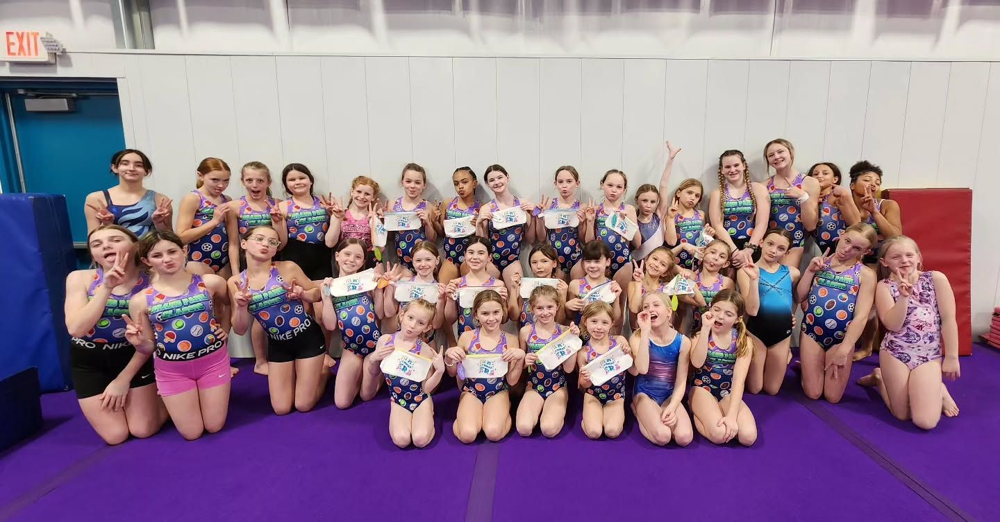 Large group posing in matching blue-purple outfits on a purple gym mat, smiling in a studio.