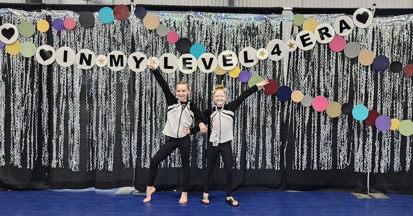 Two children standing on a stage in front of a “RUN MY LEVEL” banner and colorful garland backdrop