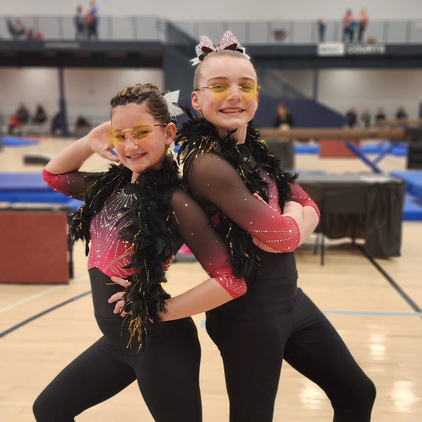 Two dancers in black and pink costumes pose back-to-back in a gymnasium, smiling with yellow glasses.