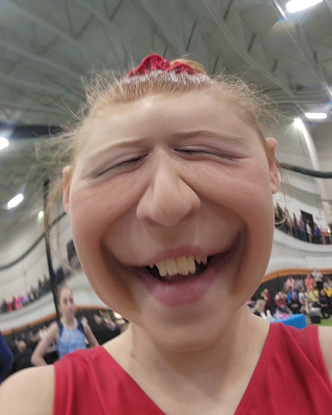 Person with a playful distorted face in a gymnasium, wearing a red top and red hair bow.