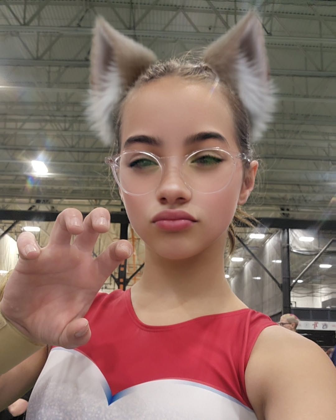 Person in a red top with a cat-ear filter, posing with a raised hand in an indoor convention hall.