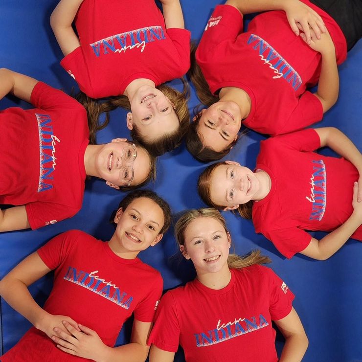 Six people in red shirts lie in a circle on a blue background, smiling up at the camera.