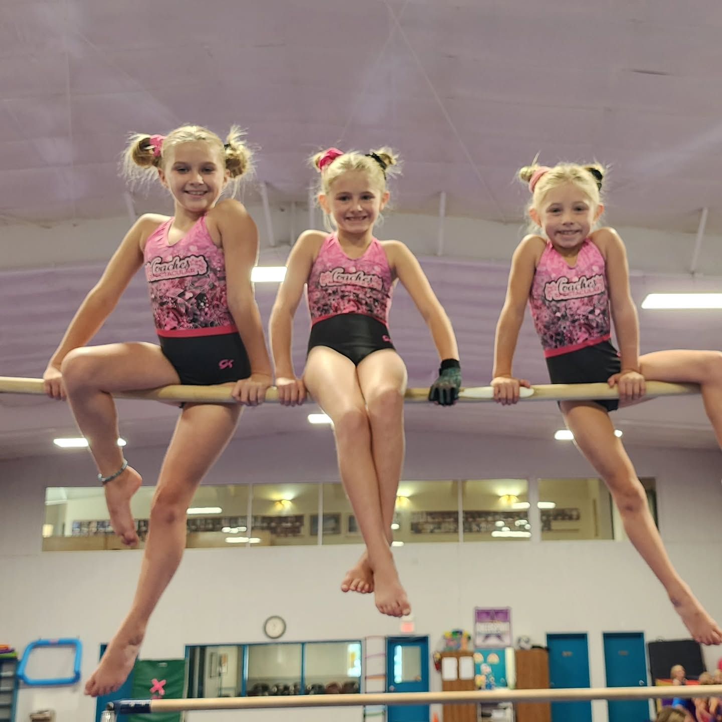 Three young gymnasts in pink leotards hang from a high bar in a gym, smiling.
