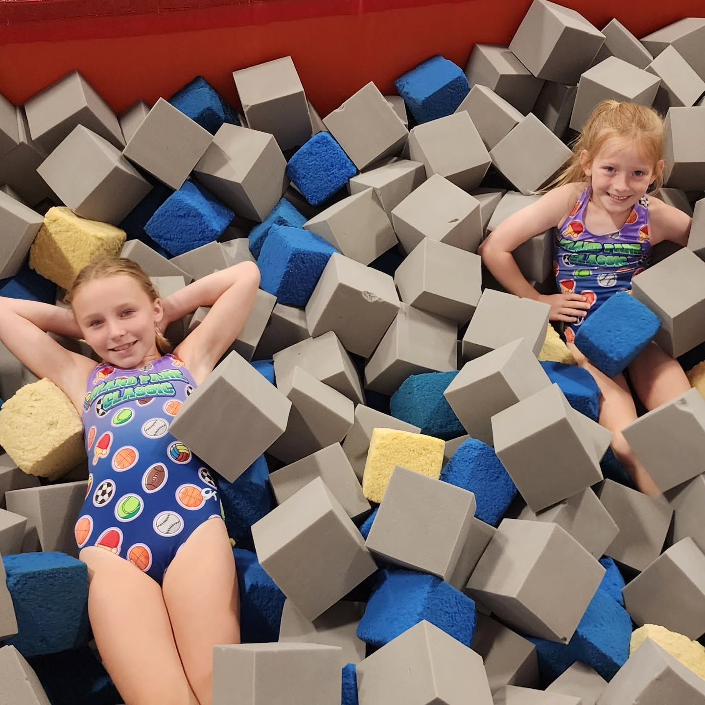 Two children lounging in a foam pit with gray and blue cubes, smiling and relaxed.