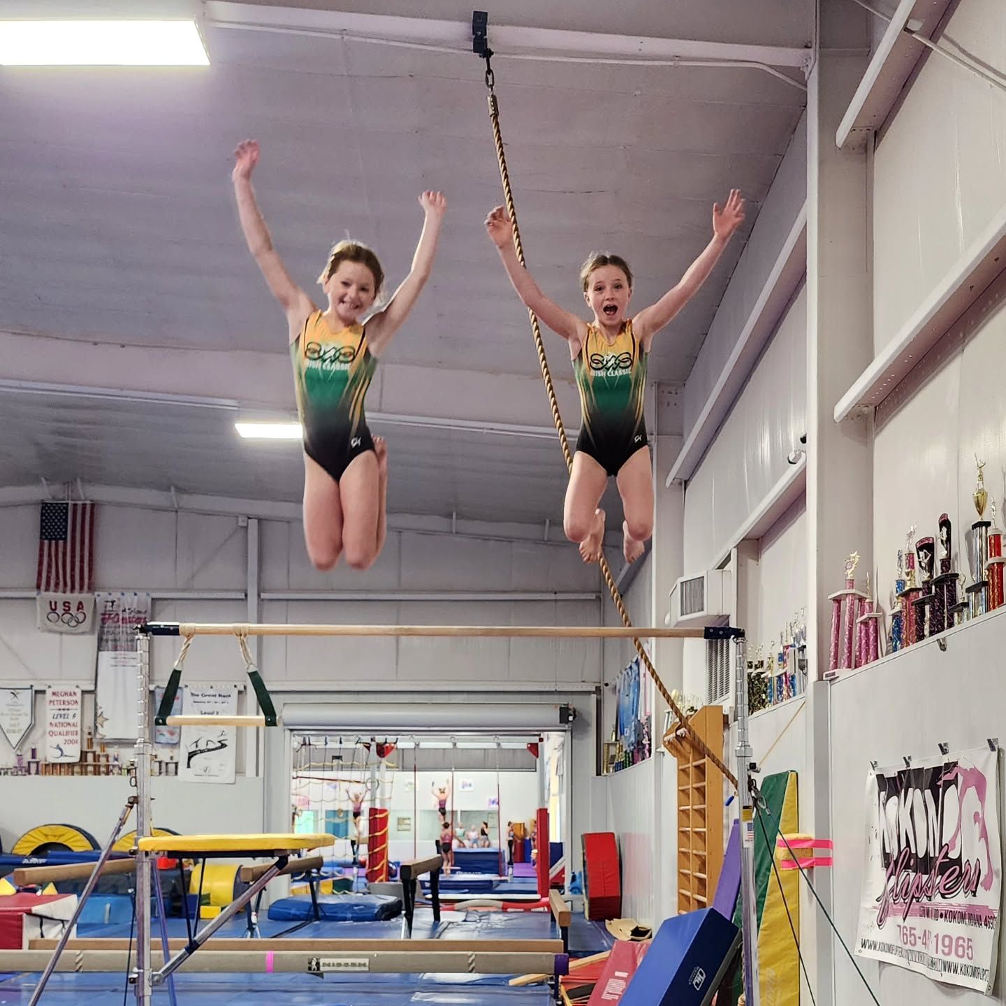 Two gymnasts in matching leotards are midair above a trampoline in an indoor gym.