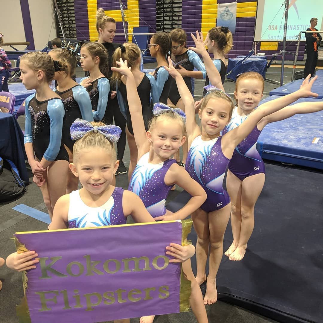 Gymnasts in blue and purple leotards hold a Kokomo Flipsters sign in a gym.