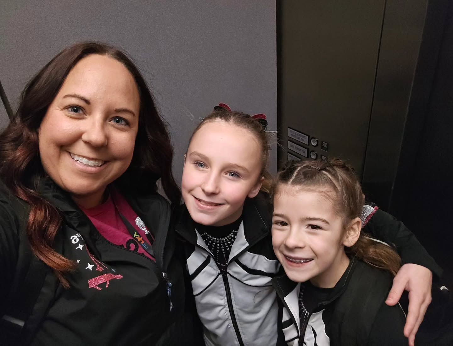 Three smiling people posing together indoors, wearing black-and-gray outfits.