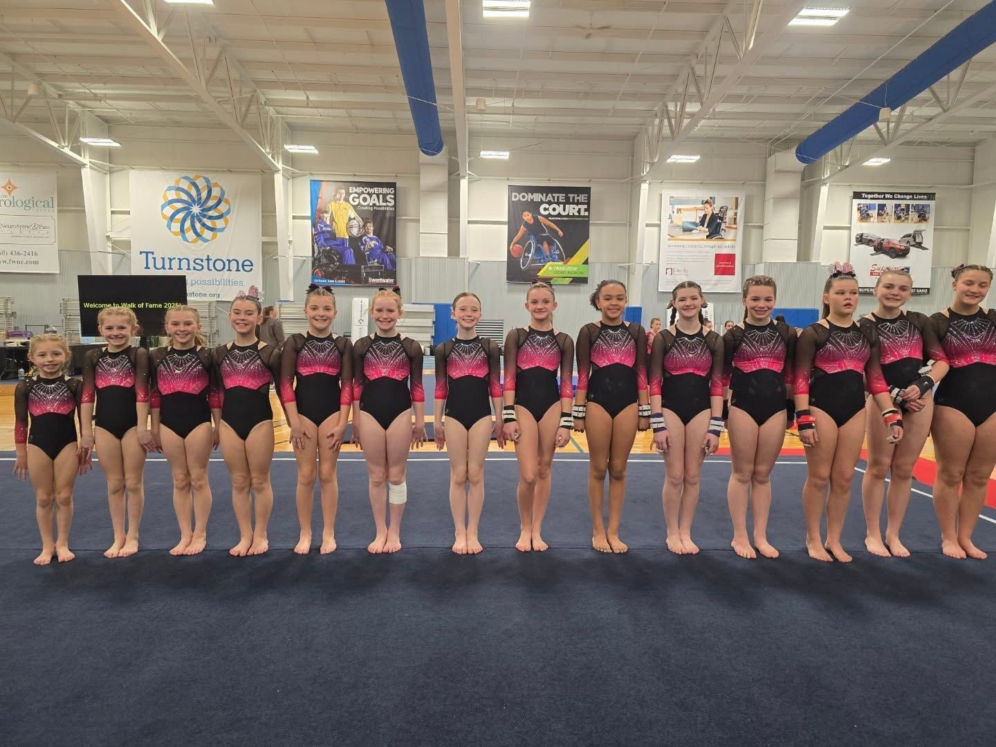 Cheer team in black and pink uniforms posing in a gym with trophies on the wall