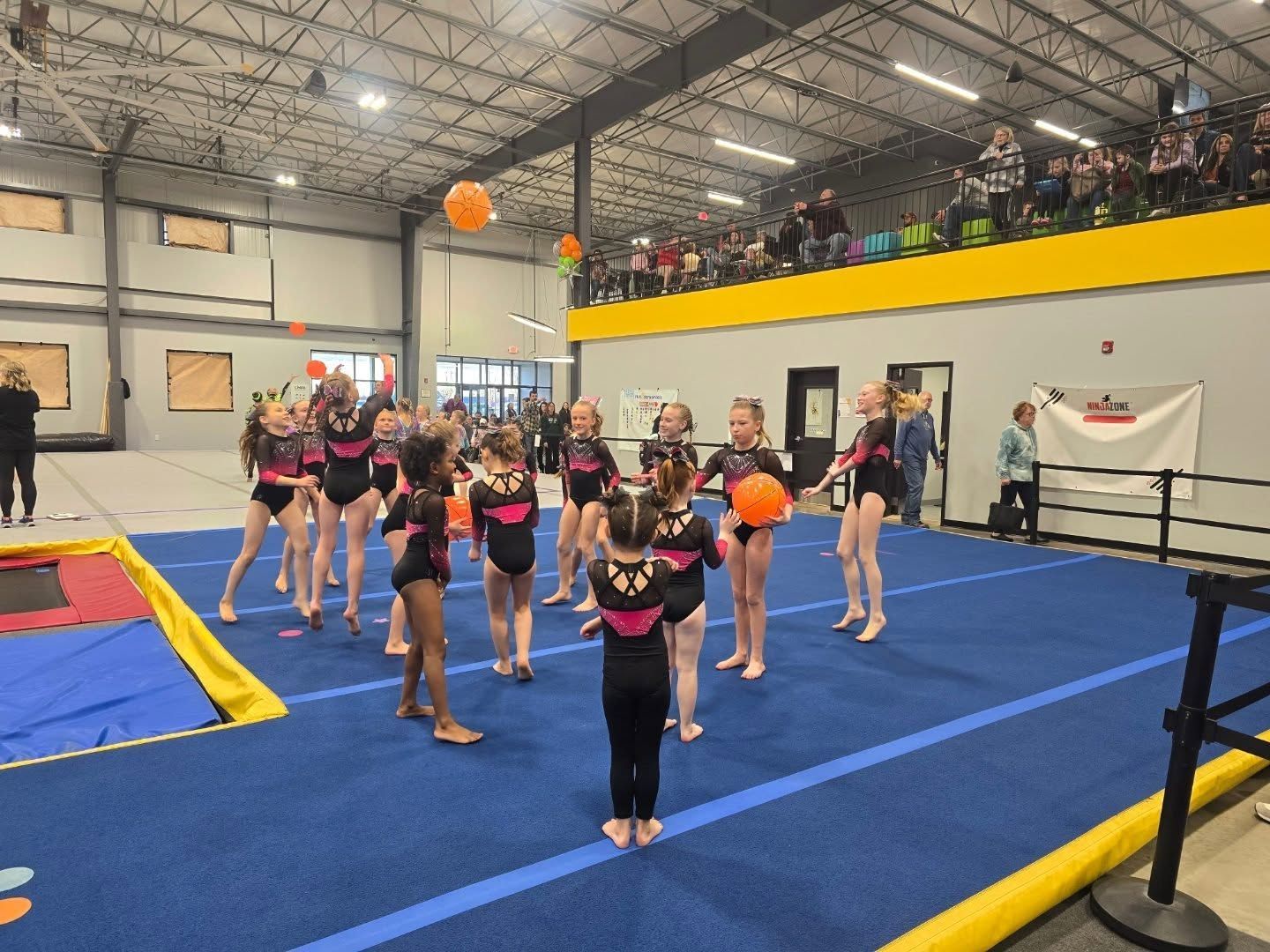 Cheerleading team performing indoors on blue mats, with orange pom-poms and spectators in a balcony above