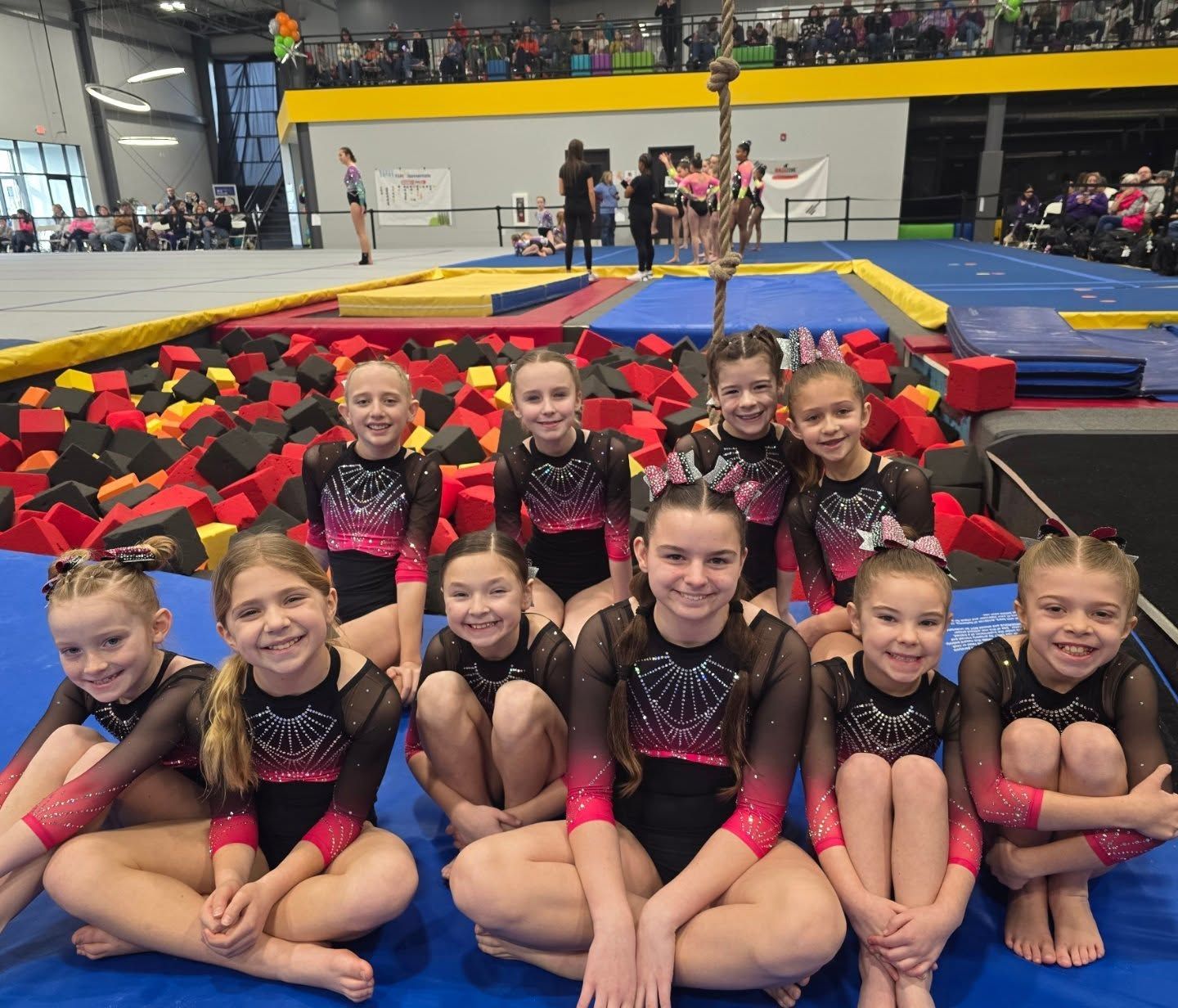Young gymnasts posing in pink and black leotards on a mat in an indoor gym arena
