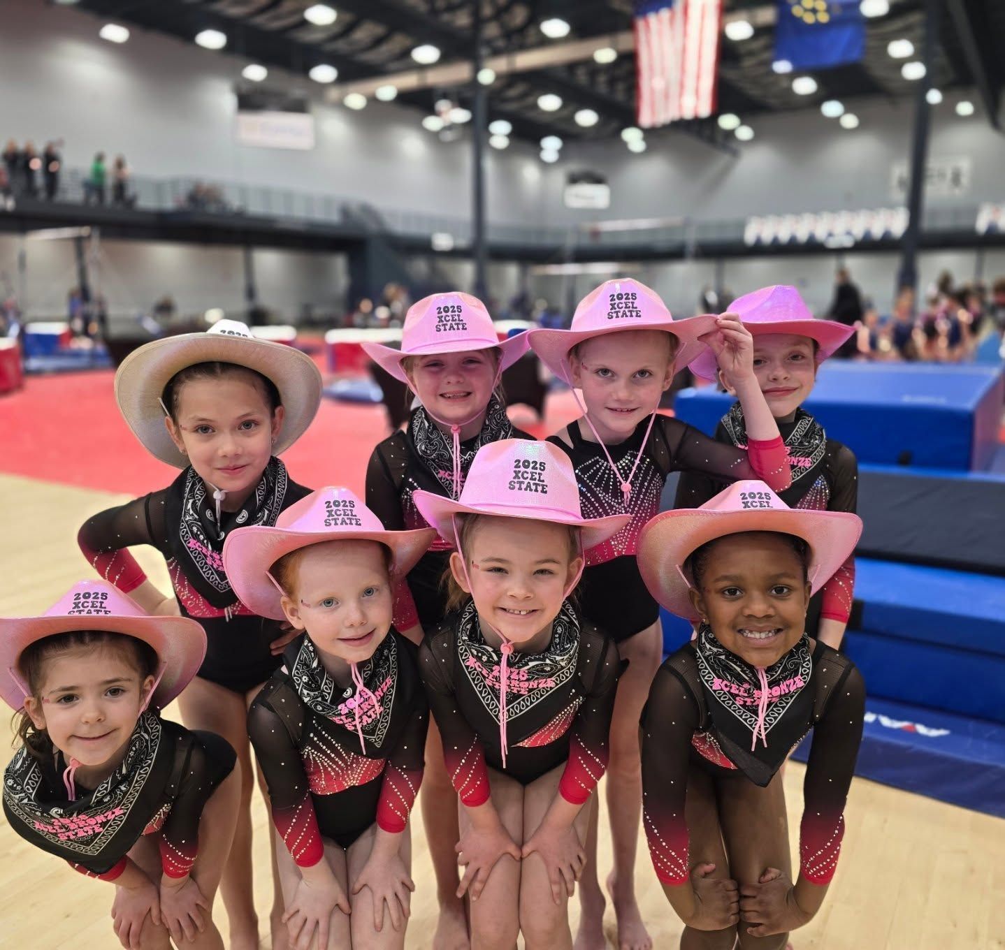 Seven cheerleaders in pink hats and black sparkly uniforms pose on a gym floor at a competition.