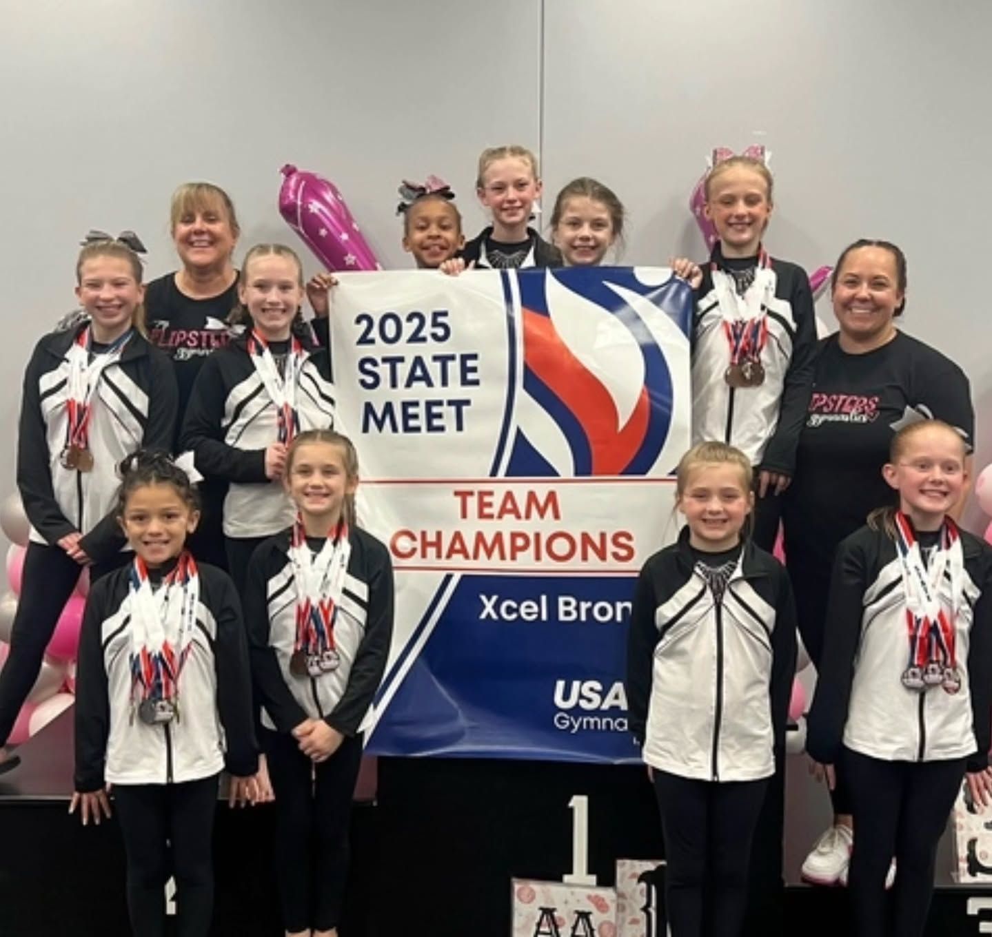 Youth gymnastics team holding “2025 State Meet Team Champions” sign on a podium, smiling in black-and-white uniforms