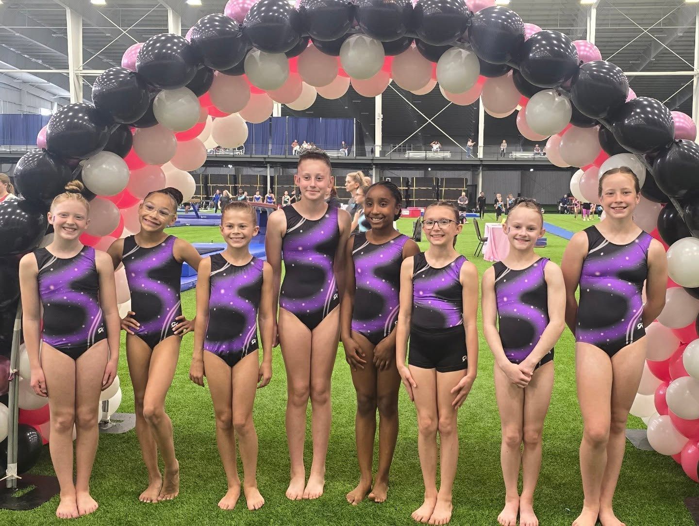 Eight gymnasts in purple-and-black leotards pose under a balloon arch at an indoor event.