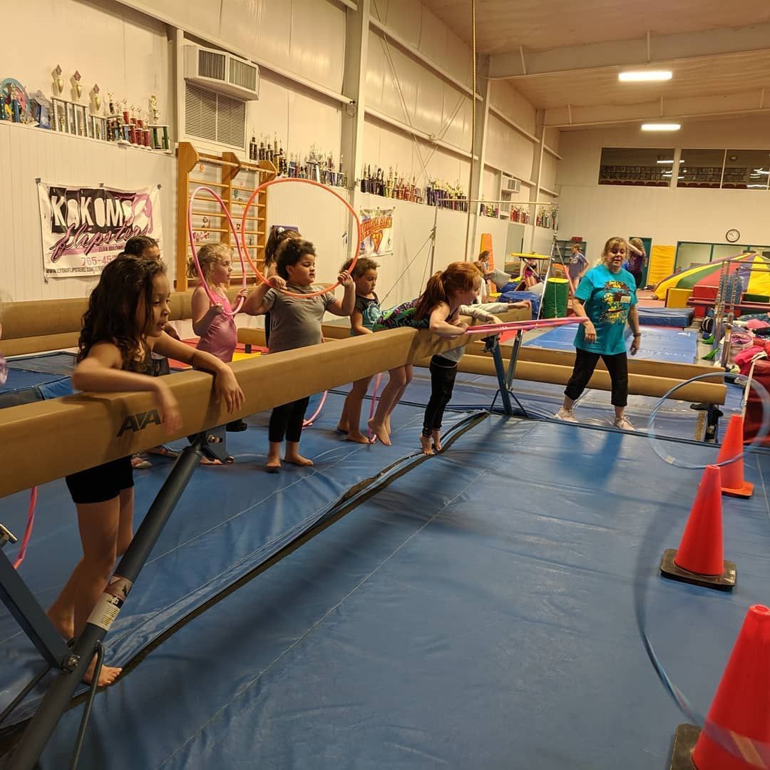 Children balancing on a wooden beam in a gym, guided by an instructor near orange cones.