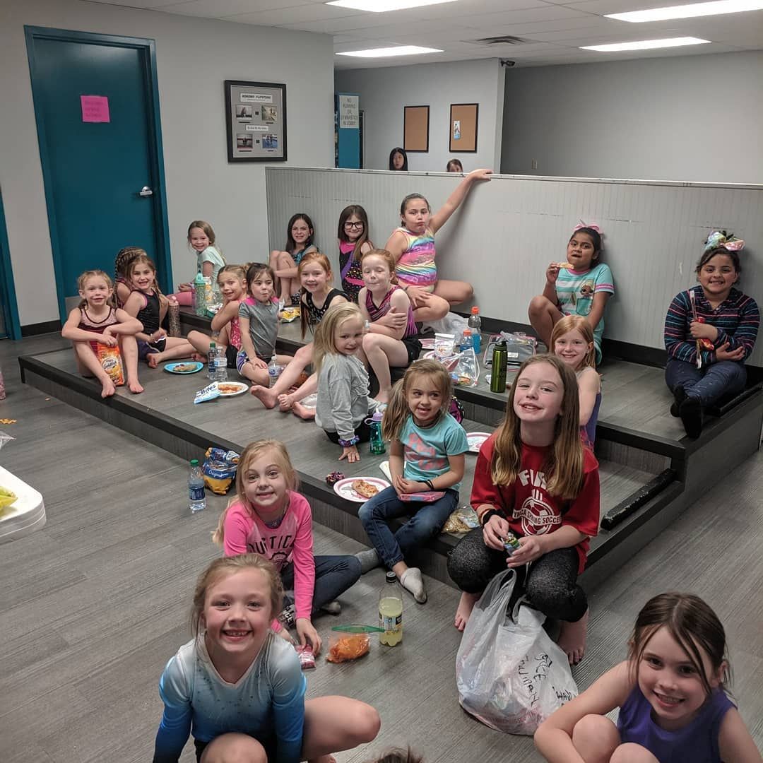 Children sitting and smiling on a raised platform in a bright indoor room, with a teal door and gray walls.