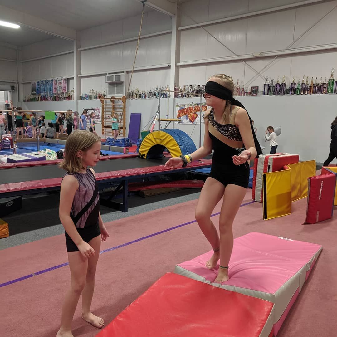 Two gymnasts on mats in a gymnastics gym, one balancing on a red beam and another standing nearby.