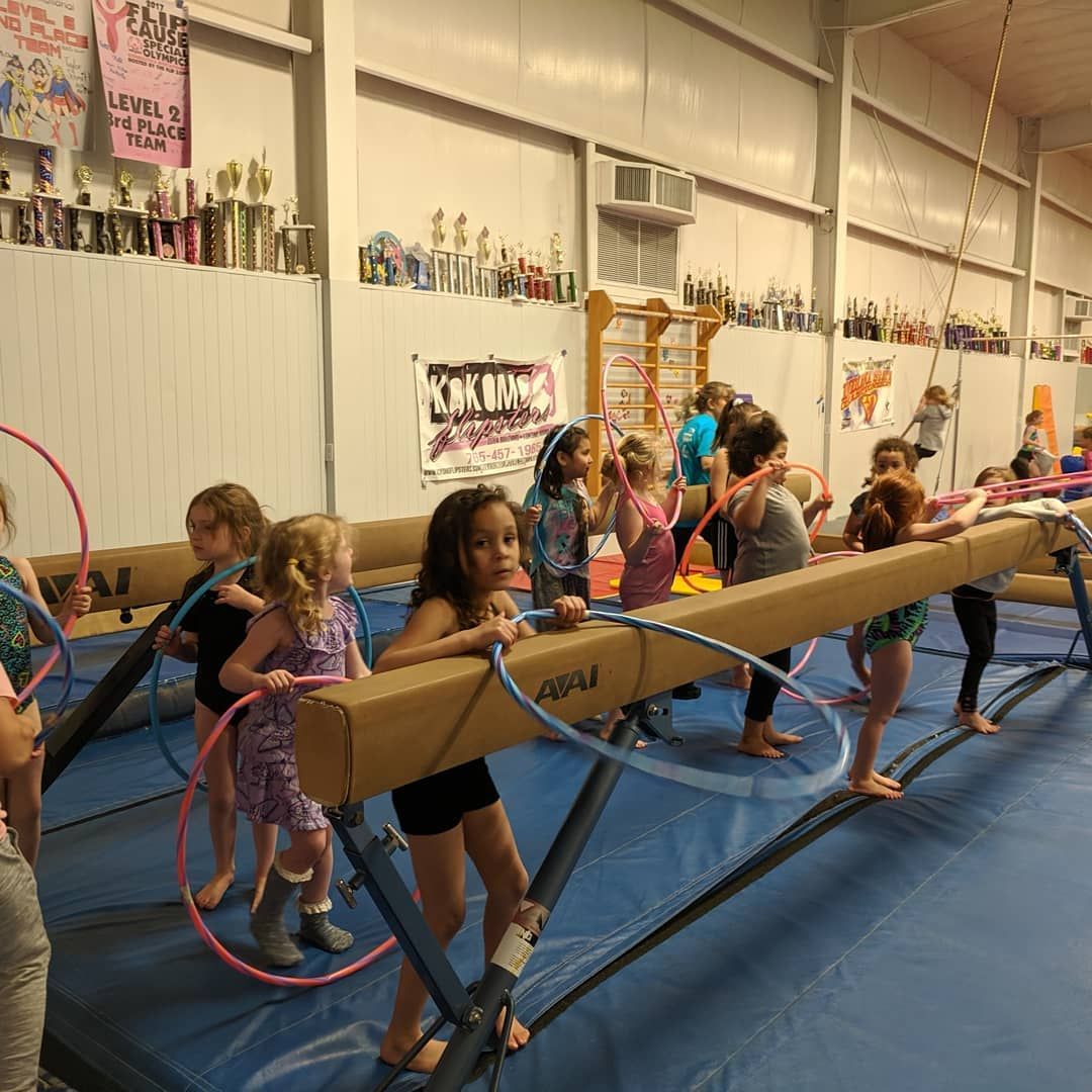 Children practicing on balance beams in a gym, with hula hoops and blue mats.