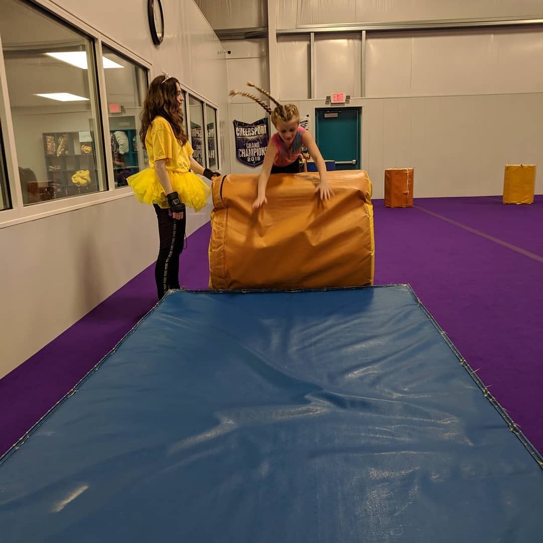 Two people in a gym beside a blue mat, holding a large orange padded block.