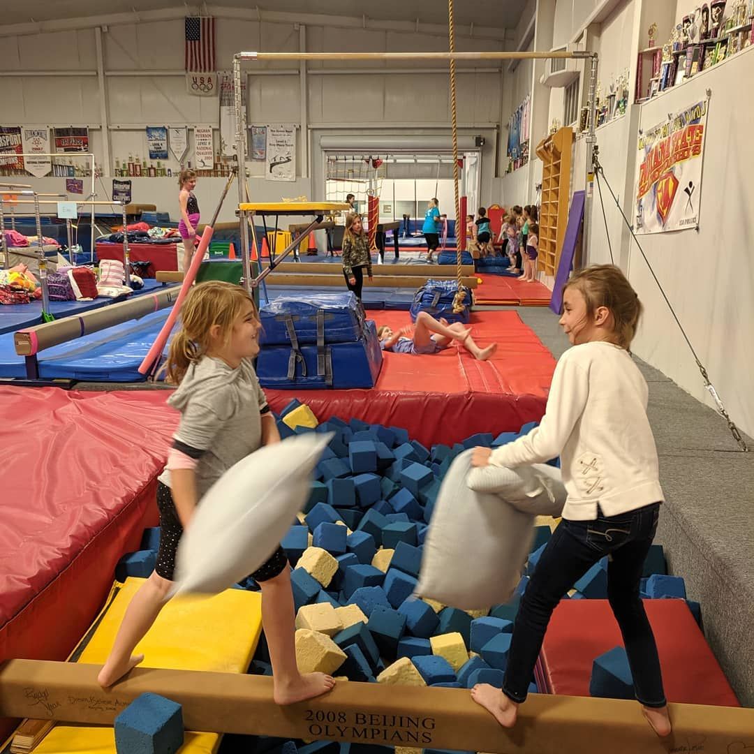 Two people sparring with foam pillows in a gym with blue foam pit and red mats.
