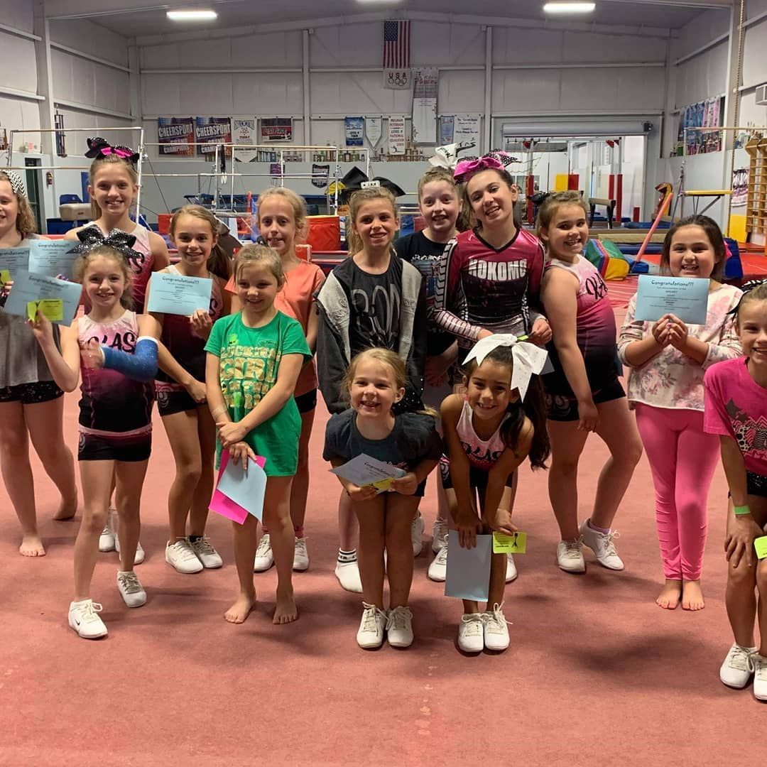 Group of children in a cheer gym, posing in colorful outfits and bows on a red mat.